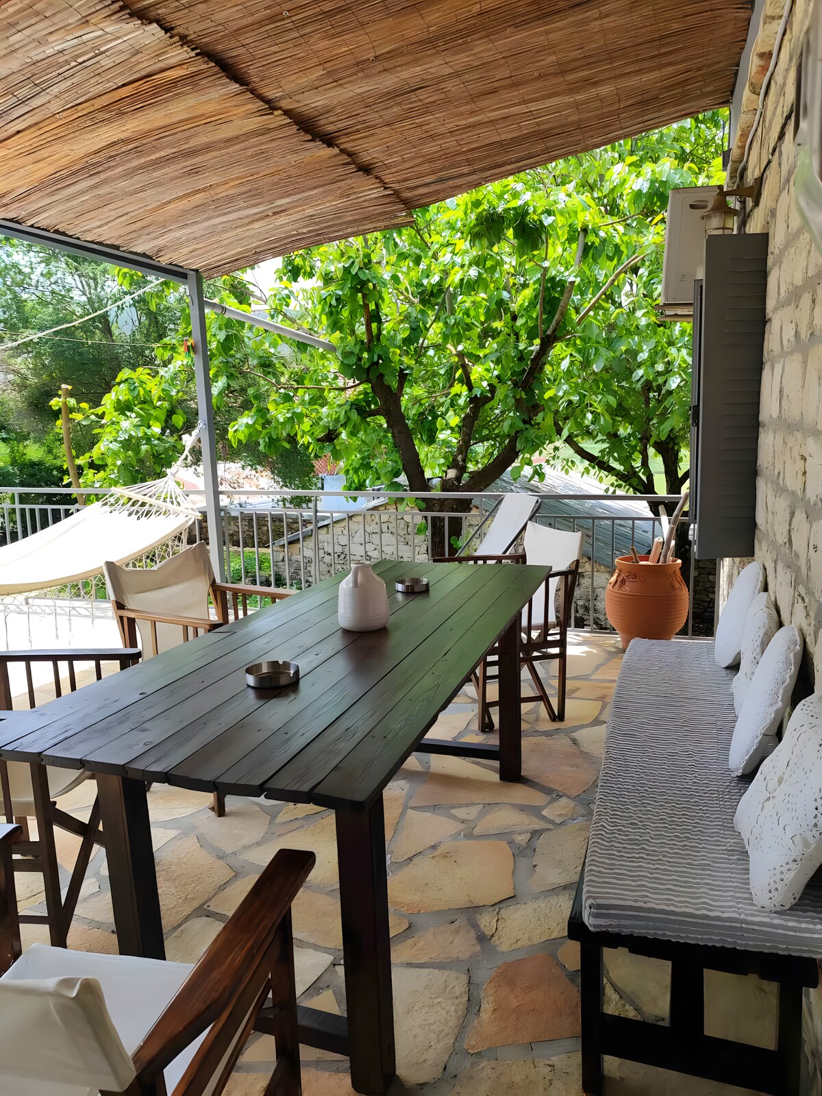 An outdoor lounge area features a wooden table surrounded by white cushioned seating. A hammock hangs nearby under a thatched roof, with lush greenery visible in the background. Natural stone flooring creates a rustic ambiance, ideal for relaxing or enjoying outdoor meals.