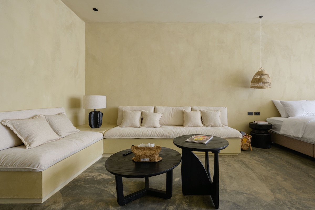 A spacious living area is adorned with soft, neutral tones, featuring two large built-in sofas with plush cushions. A round black coffee table sits at the center, accompanied by a bedside table and a stylish lamp. Natural light filters in, enhancing the serene ambiance.