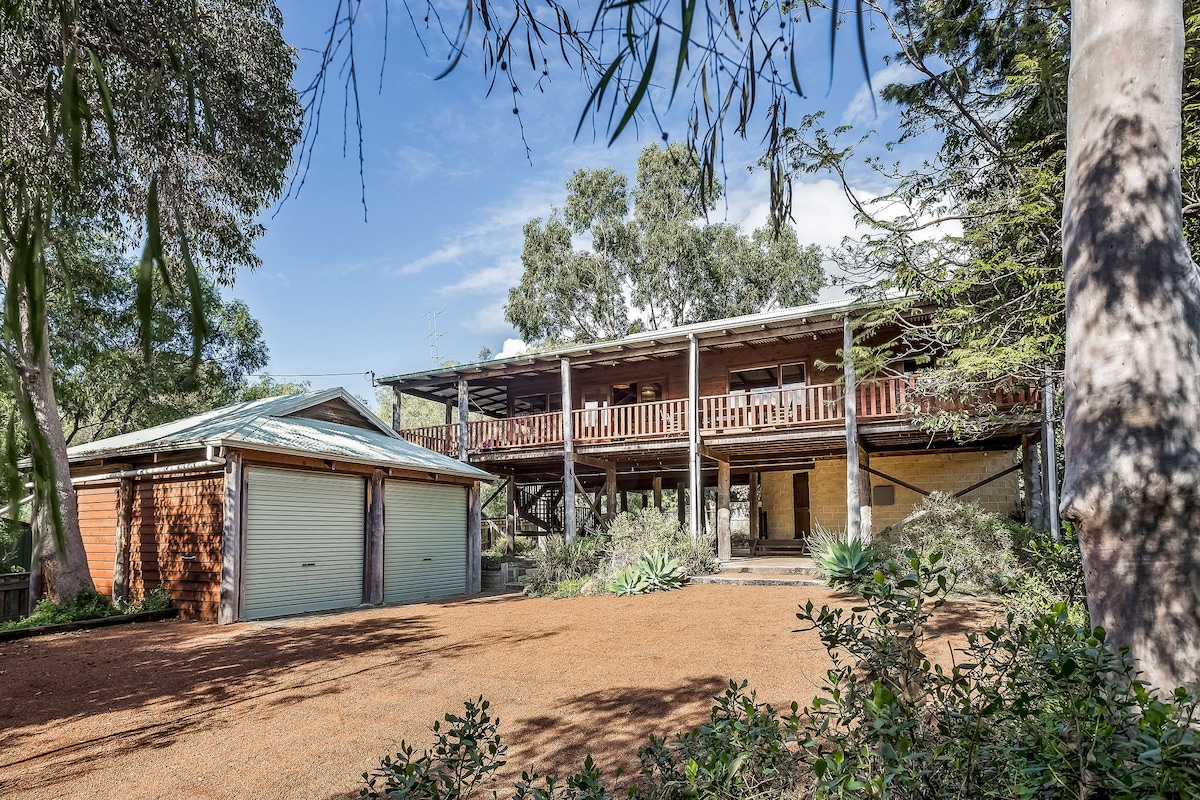 A two-story wooden home is nestled among trees, featuring a spacious wrap-around balcony with wooden railings. A separate garage building is visible to the left. The driveway is lined with gravel and surrounded by native plants, enhancing the peaceful coastal setting.