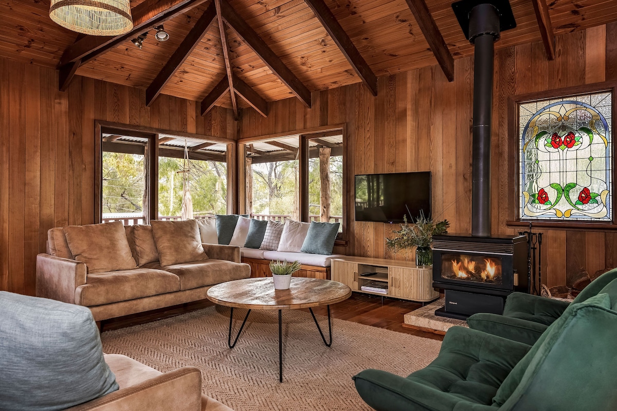 A cozy living area is showcased with wooden walls and a high-beamed ceiling. A large round coffee table sits in the center, surrounded by comfortable sofas and armchairs. A wood-burning stove adds warmth, while stained glass windows provide colorful accents and natural light.