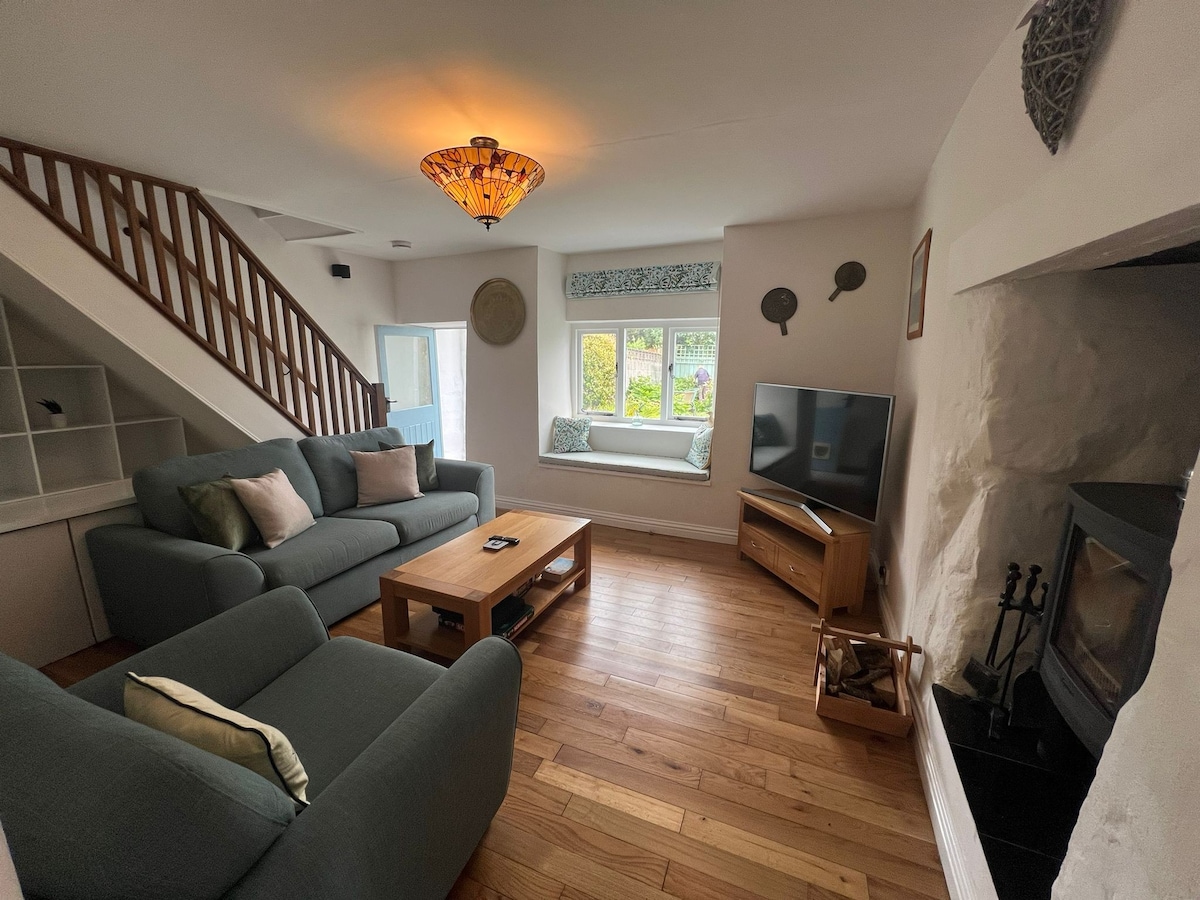 A cozy sitting room features comfortable sofas arranged around a wooden coffee table. A television is positioned on an adjacent cabinet. Light wood flooring is complemented by a warm wooden staircase. A wood burner is situated in the corner, with natural light entering through windows adorned with fabric treatments.