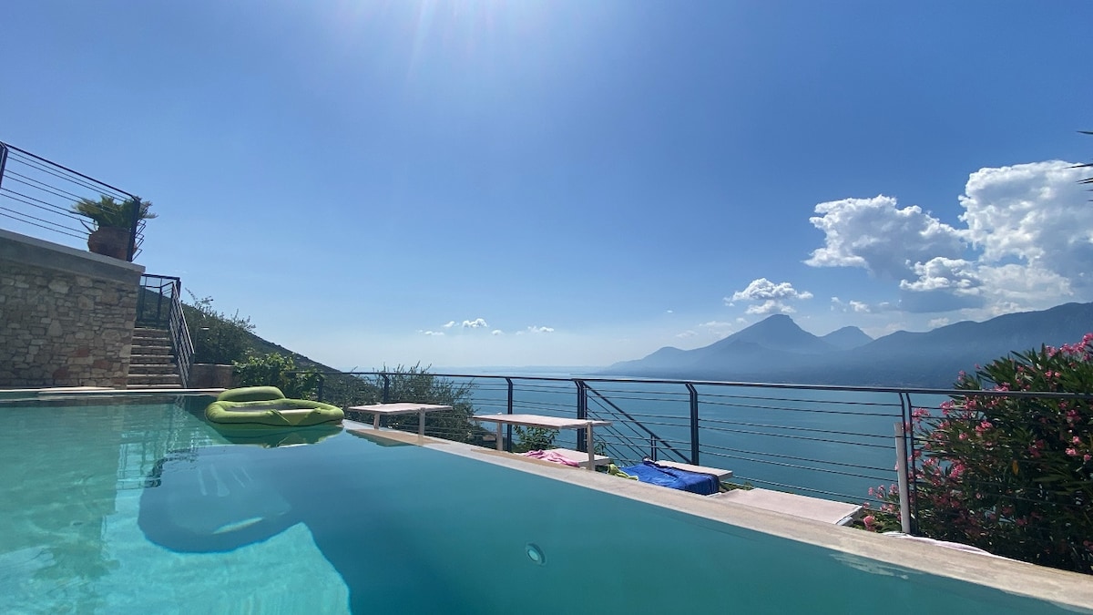 A serene pool area is visible, featuring calm waters reflecting the blue sky. Lush greenery and colorful lounge chairs border the pool. In the distance, rolling hills and mountains rise above the tranquil sea, creating a peaceful backdrop under soft, fluffy clouds.