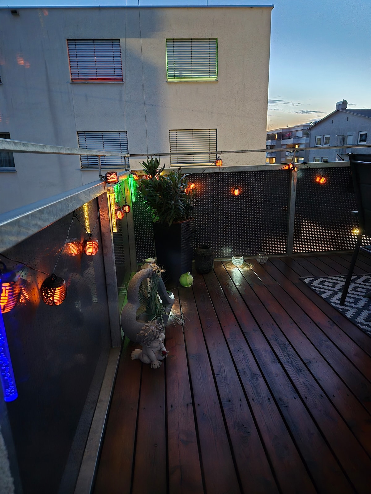 A balcony space features wooden decking adorned with string lights and decorative lanterns. A potted plant is positioned near the corner, while a small decorative figure adds character. The scene captures an inviting atmosphere during dusk, with city buildings visible in the background.