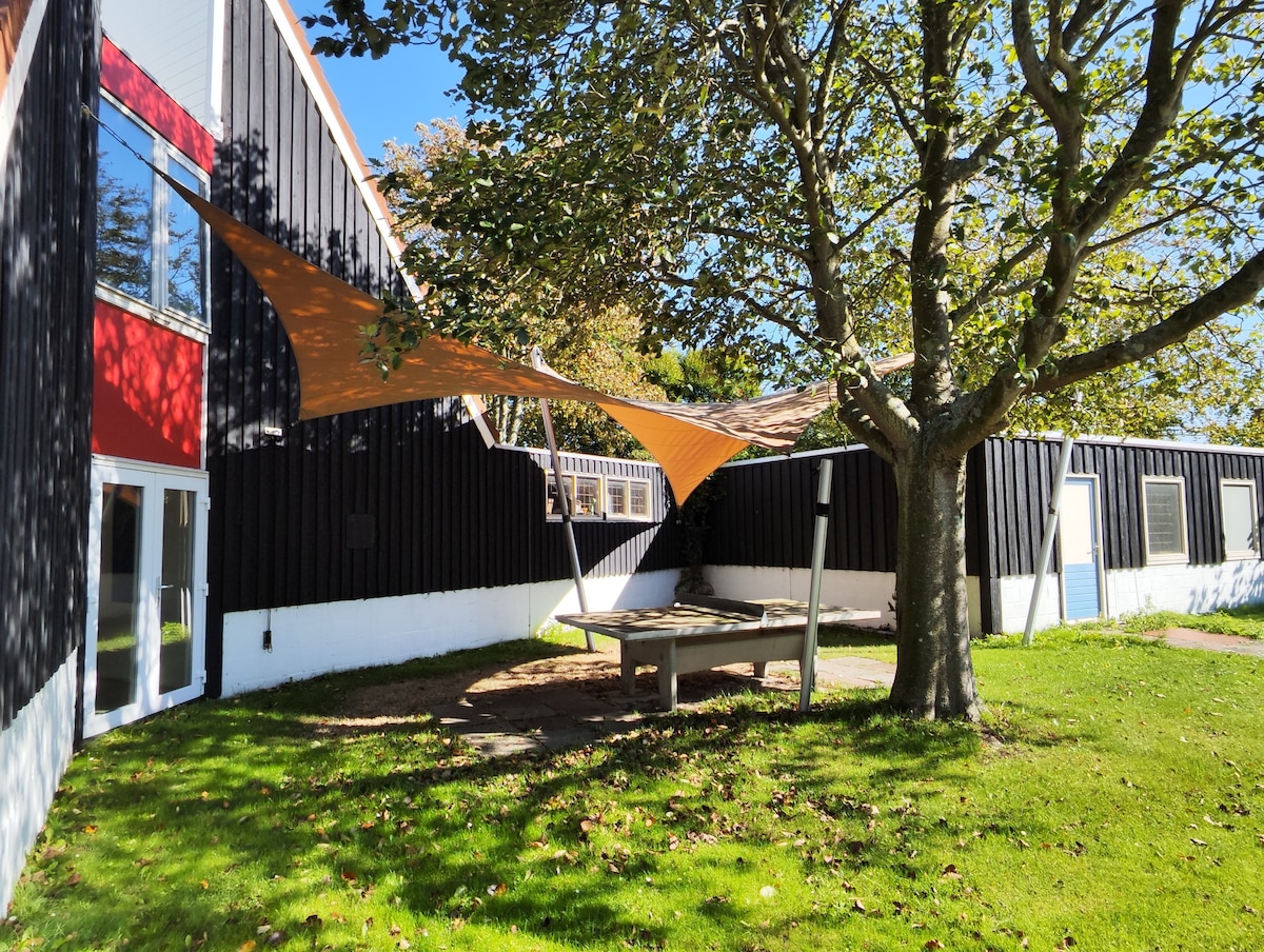An outdoor area is featured, surrounded by lush green grass and shaded by a large tree. A sunshade covers a picnic table, providing protection from the sun. The building's modern design includes a mix of black and red panels, contributing to a welcoming atmosphere.