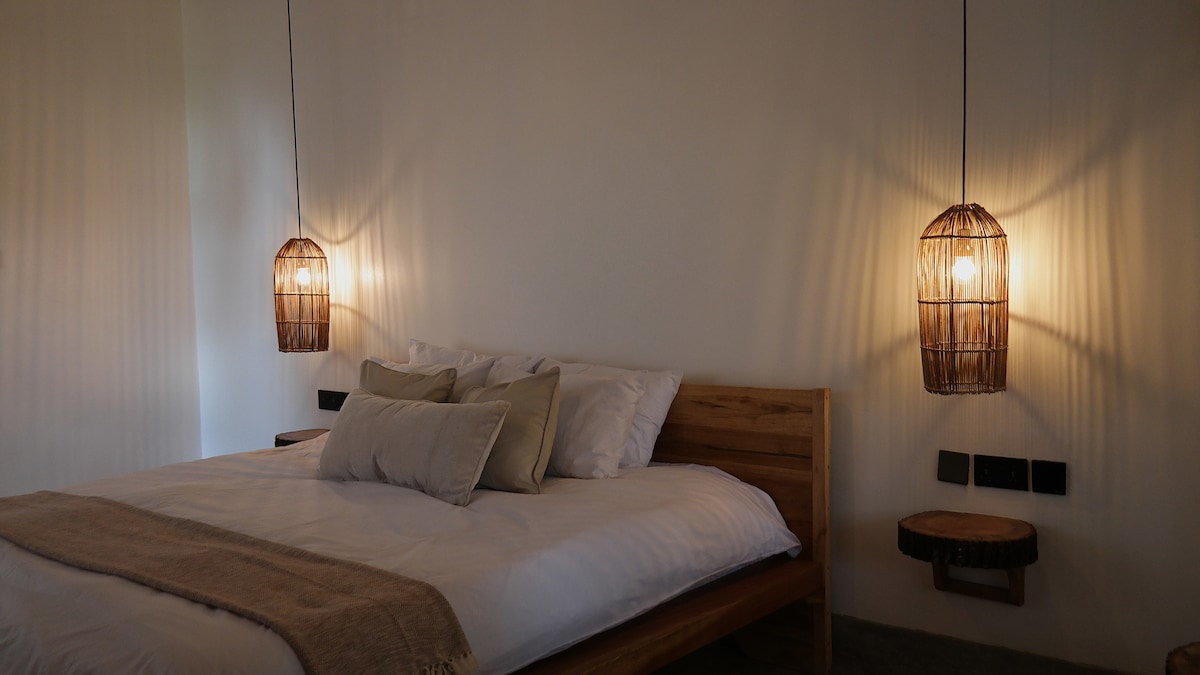 A serene bedroom scene is presented with a wooden bed topped by plush pillows and a soft blanket. Two woven pendant lights hang above, providing warm illumination. The walls are painted in a soft hue, enhancing the calm ambiance of the space.