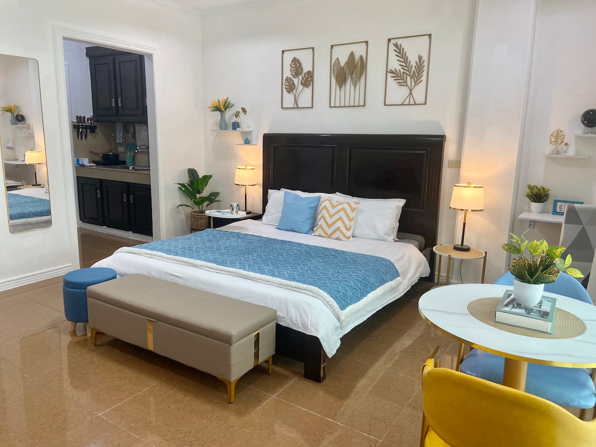 The studio apartment features a spacious bed with a light blue and white bedspread. A sleek kitchenette is visible in the background, complemented by modern furniture. Decorative wall art enhances the bright atmosphere, while a dining table with colorful chairs is positioned to the right.