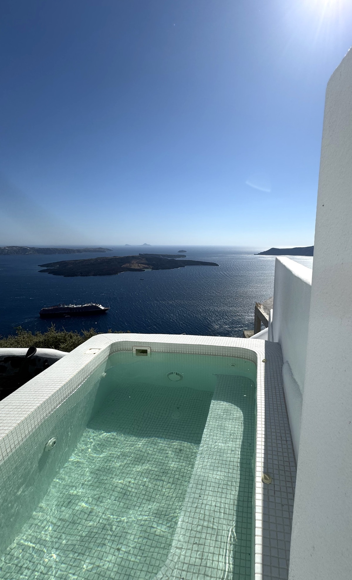 An outdoor jacuzzi is displayed on a private terrace, offering panoramic views of the Aegean Sea. The clear blue water reflects sunlight, enhancing the serene ambiance. The coastline and distant islands are visible, contributing to a sense of tranquility.