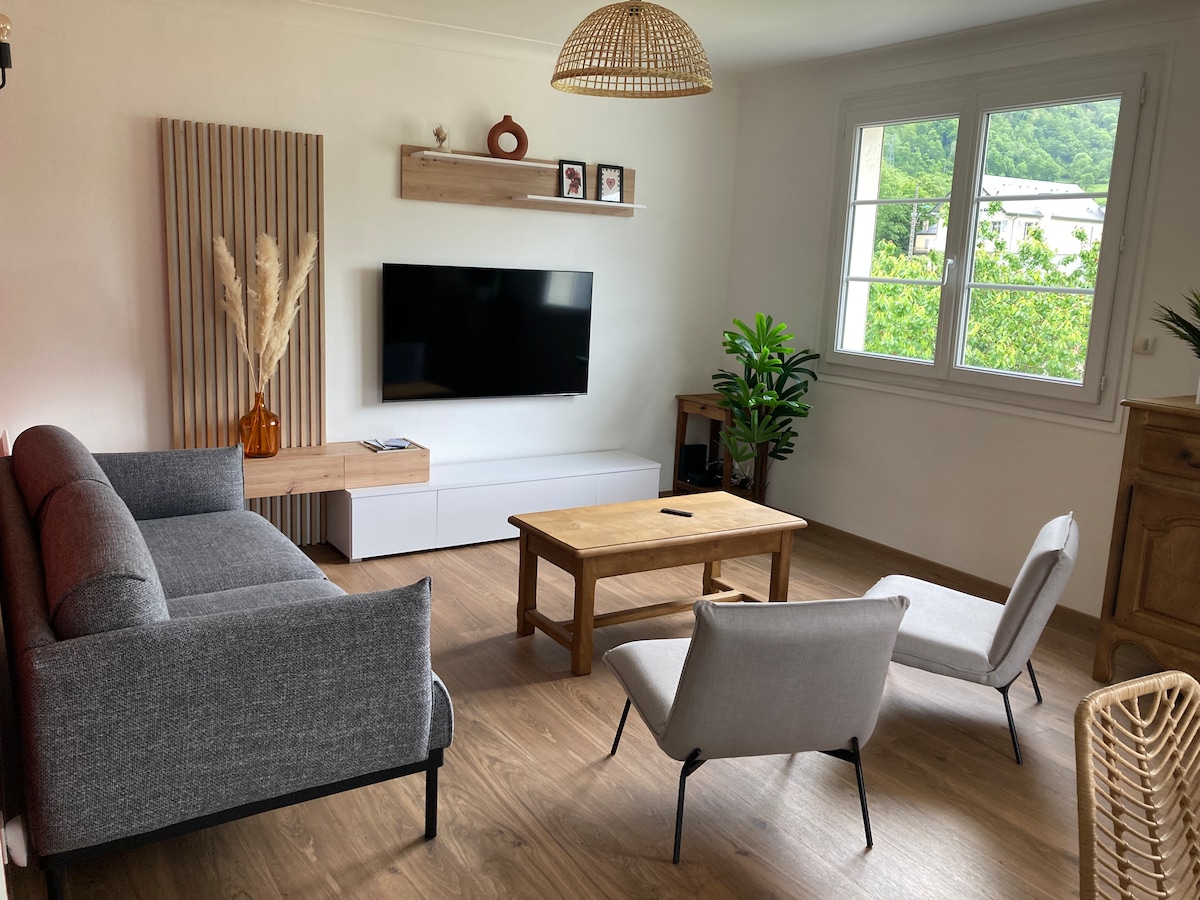 A living area is presented with a comfortable grey sofa and two light chairs. A wooden coffee table sits at the center, complemented by a flat-screen TV mounted on the wall. Bright light enters through the window, enhancing the greenery visible outside.