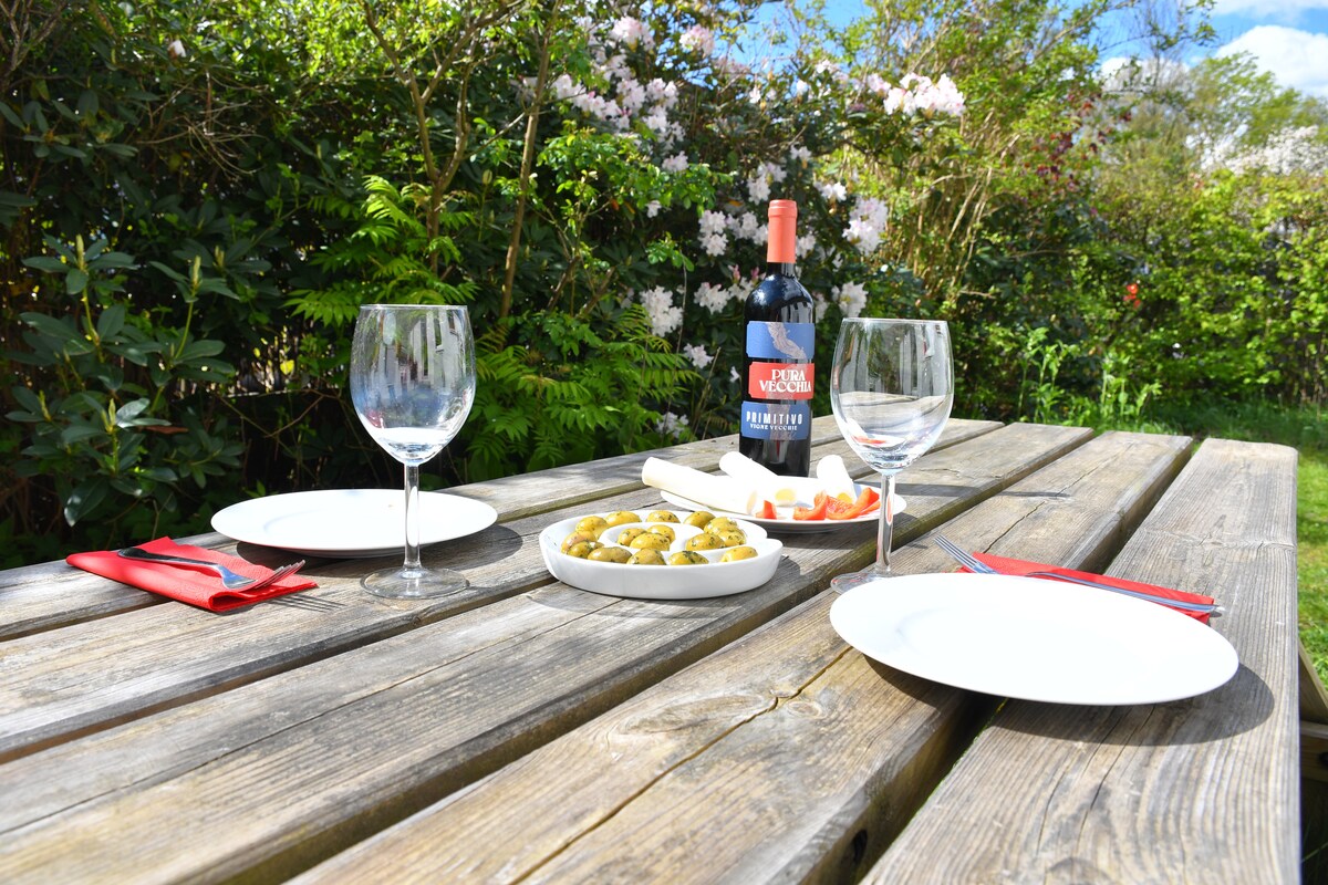 A wooden picnic table is set for a meal in a lush garden. Two wine glasses sit beside three plates, accompanied by a platter of olives and fresh tomatoes. A bottle of wine is positioned at the center, surrounded by vibrant greenery and blooming flowers in the background.