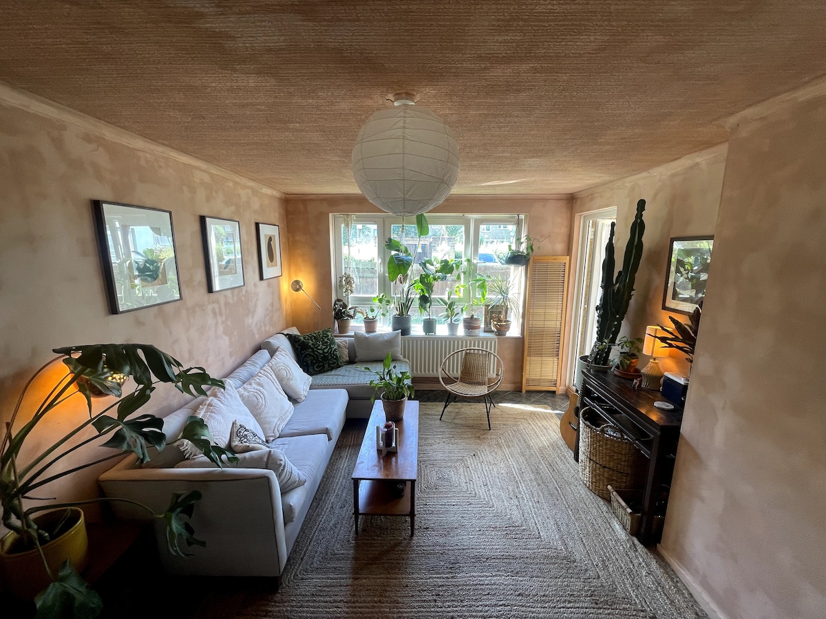 A bright living space is filled with natural light, featuring a comfortable sofa adorned with cushions. Lush green plants are positioned near large windows, while a wooden coffee table and a woven rug complete the cozy ambiance. Art hangs on the textured walls, enhancing the serene environment.