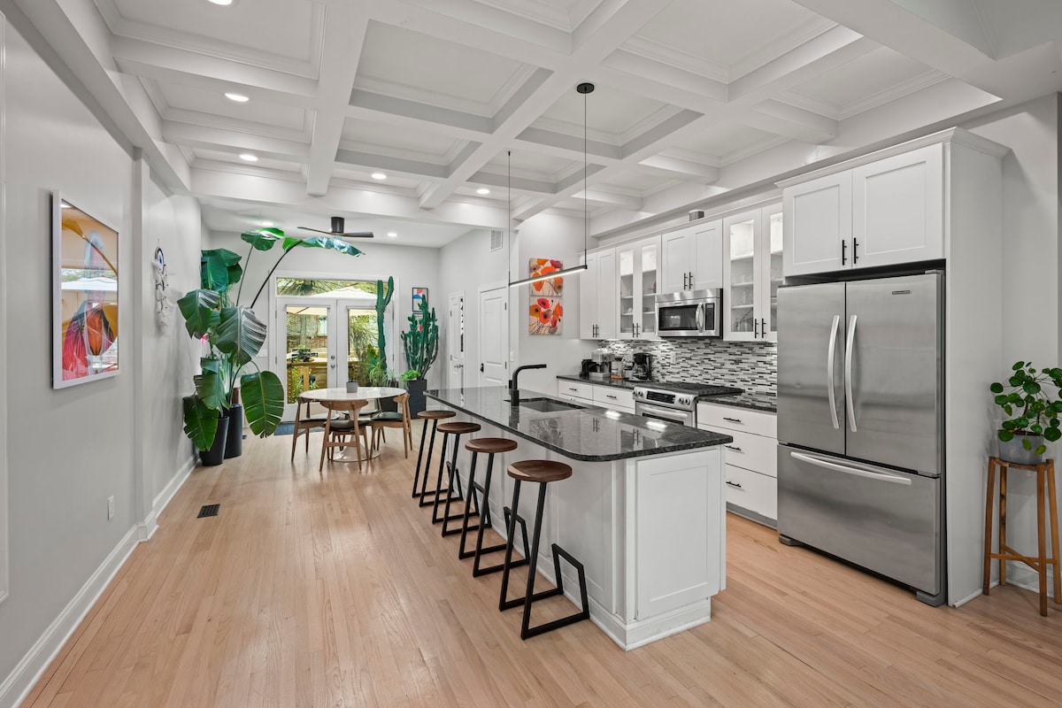 A modern kitchen is characterized by white cabinetry and a large island topped with dark granite. Sleek appliances are visible, including a stainless steel refrigerator and microwave. A row of wooden stools accompanies the island, while greenery and artwork add vibrant touches to the airy space.