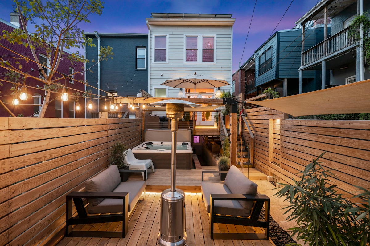 A spacious outdoor oasis is presented, featuring two lounge areas with modern seating and a hot tub surrounded by wooden decking. Soft lighting from string lights enhances the warm ambiance, while a patio umbrella provides shade, creating an inviting space for relaxation.