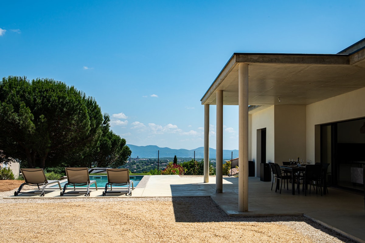 An outdoor patio area includes lounge chairs positioned by a clear pool, surrounded by a sunlit landscape. A dining table is visible nearby, providing a space for outdoor meals. The backdrop features rolling hills under a bright blue sky.