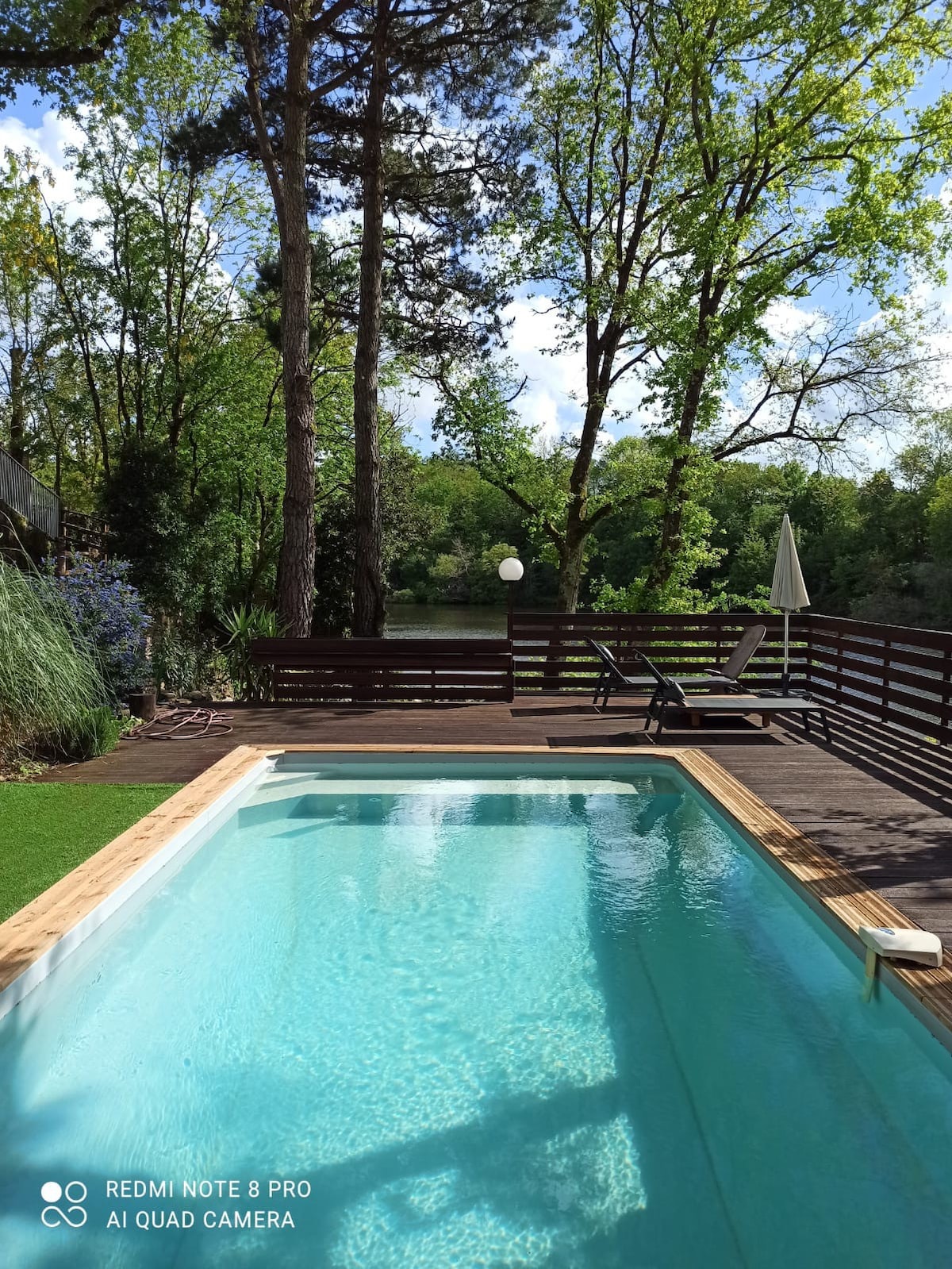 A serene swimming pool is framed by a wooden deck, offering views of lush greenery and a peaceful lake. Sun loungers are positioned nearby, and a shaded umbrella provides a relaxing spot. The area is surrounded by tall trees, enhancing the tranquil atmosphere.