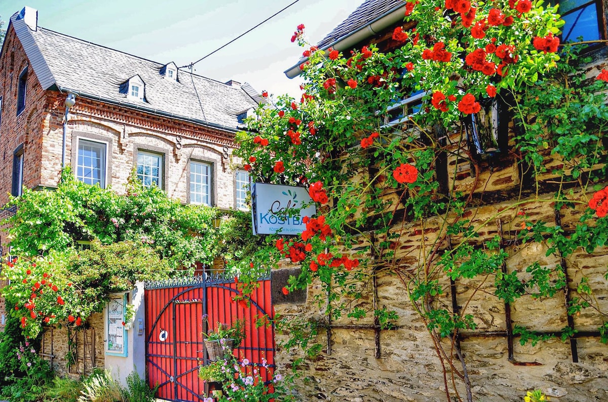 A charming exterior view features a stone wall adorned with blooming red flowers. The entrance is marked by a vibrant red gate, leading to a welcoming setting. A traditional building with a sloped roof is partially visible, surrounded by greenery.