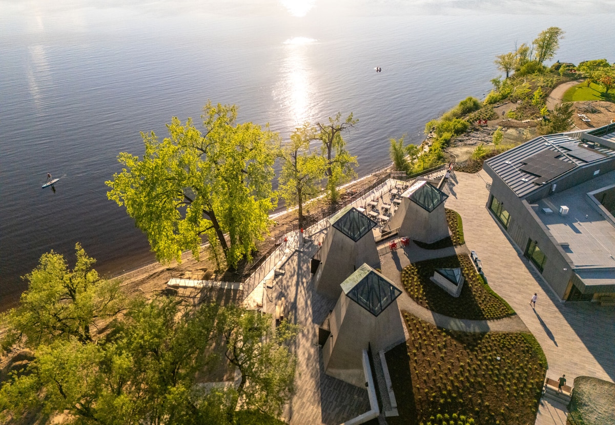 An aerial view showcases a serene lakeside landscape with gentle waves lapping at the shore. Modern structures with angular designs are nestled among lush green trees, and pathways meander toward the water. Light reflects off the lake, creating a tranquil atmosphere.