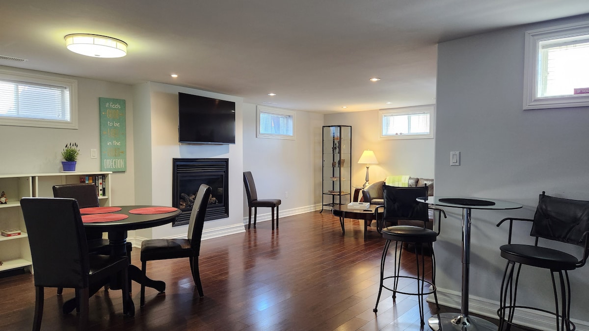 A spacious living area features warm wooden flooring and ample natural light from multiple windows. A dining table with four chairs is positioned near the wall, while a cozy seating arrangement includes a Sofa and a lamp. A mounted TV and fireplace add functional elements.