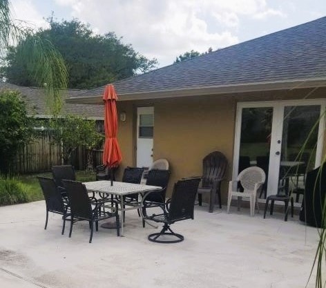 An outdoor patio area is showcased, featuring a large table surrounded by several chairs, including two swivel seats. A bright orange umbrella provides shade. The setting is complemented by greenery in the background, with a nearby door leading into the home.