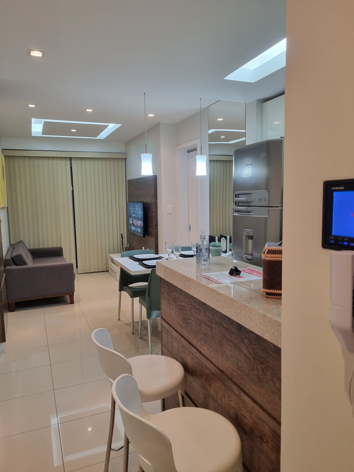 A modern kitchen area is visible, featuring sleek granite countertops and bar stools. Bright pendant lights illuminate the space. A dining table is set for two, while the living area, with a comfortable sofa, is partially seen through sliding doors. Natural light enters through a skylight.