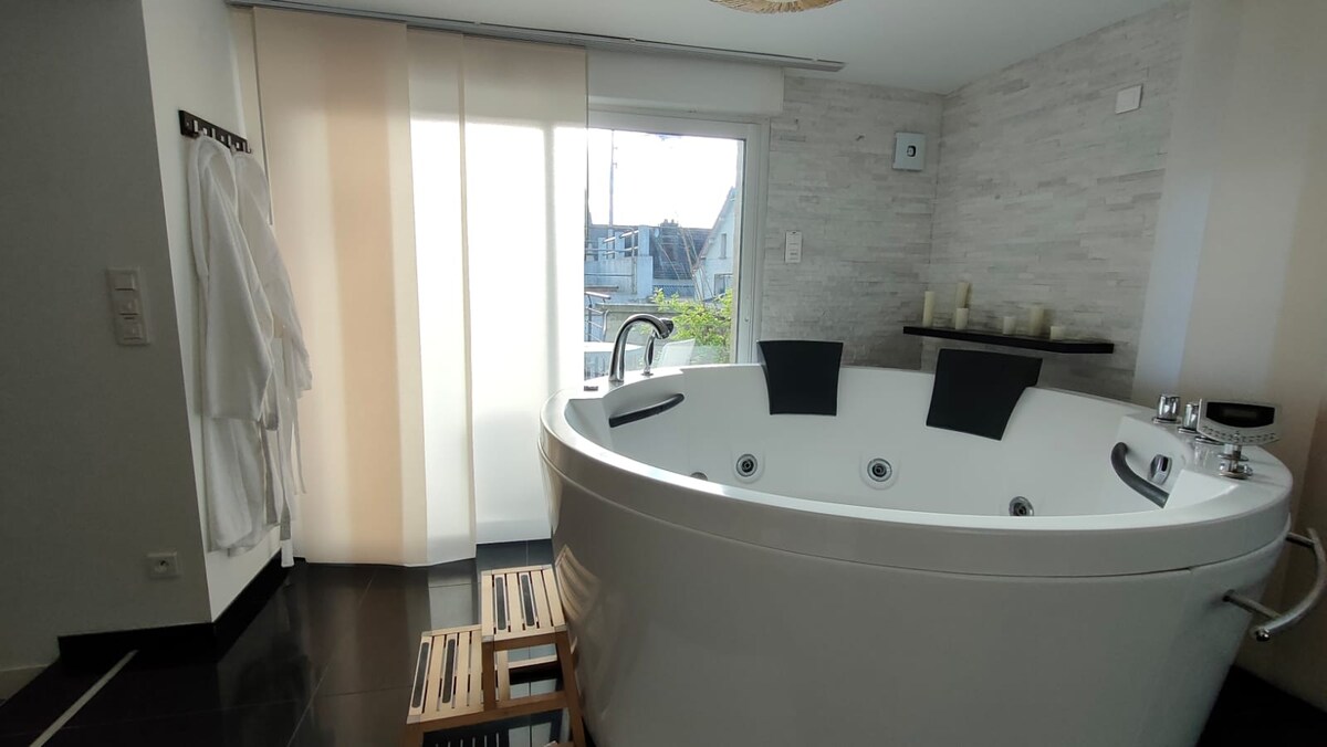 A spacious bathroom features a round balnéo bathtub with three integrated seats. Natural light enters through a partially covered window, highlighting the modern design. Soft bathrobes are hung nearby, and a wooden stool adds a touch of warmth to the tiled floor.