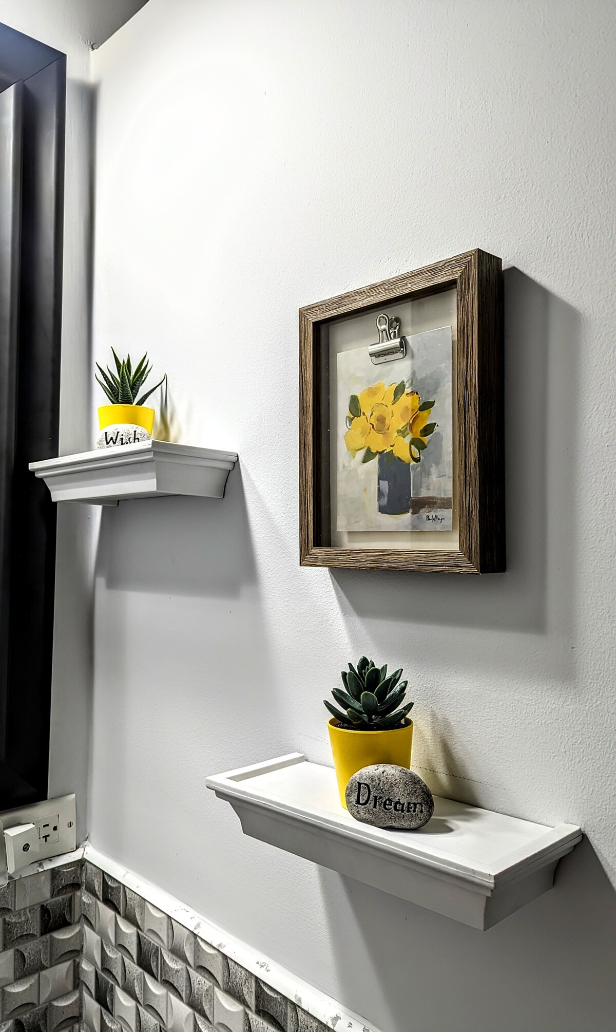 A bathroom wall features two white shelves displaying small potted plants. A framed painting of yellow flowers hangs in between, adding a touch of color. The decor emphasizes simplicity and warmth in the space.