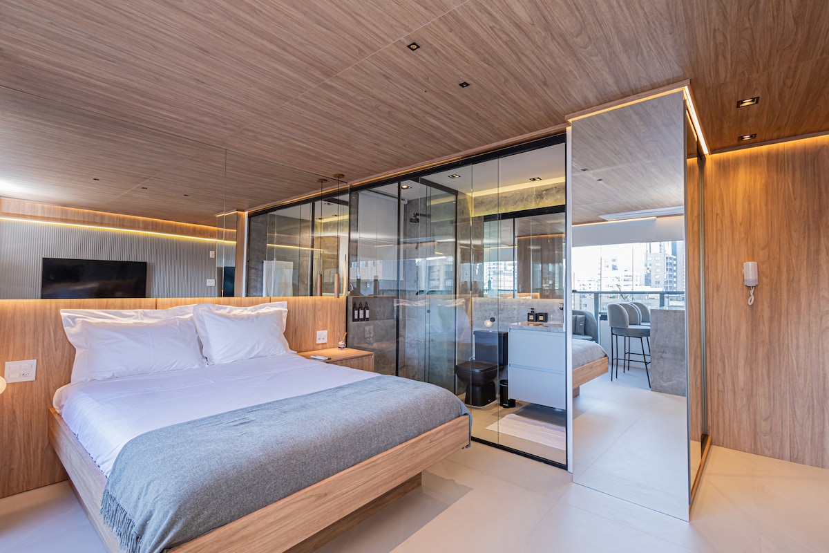 A modern studio space features a spacious bed with neutral bedding, complemented by wooden paneling and sleek glass partitions. Soft lighting highlights the area, with a wall-mounted television visible and a balcony area framed by floor-to-ceiling windows.