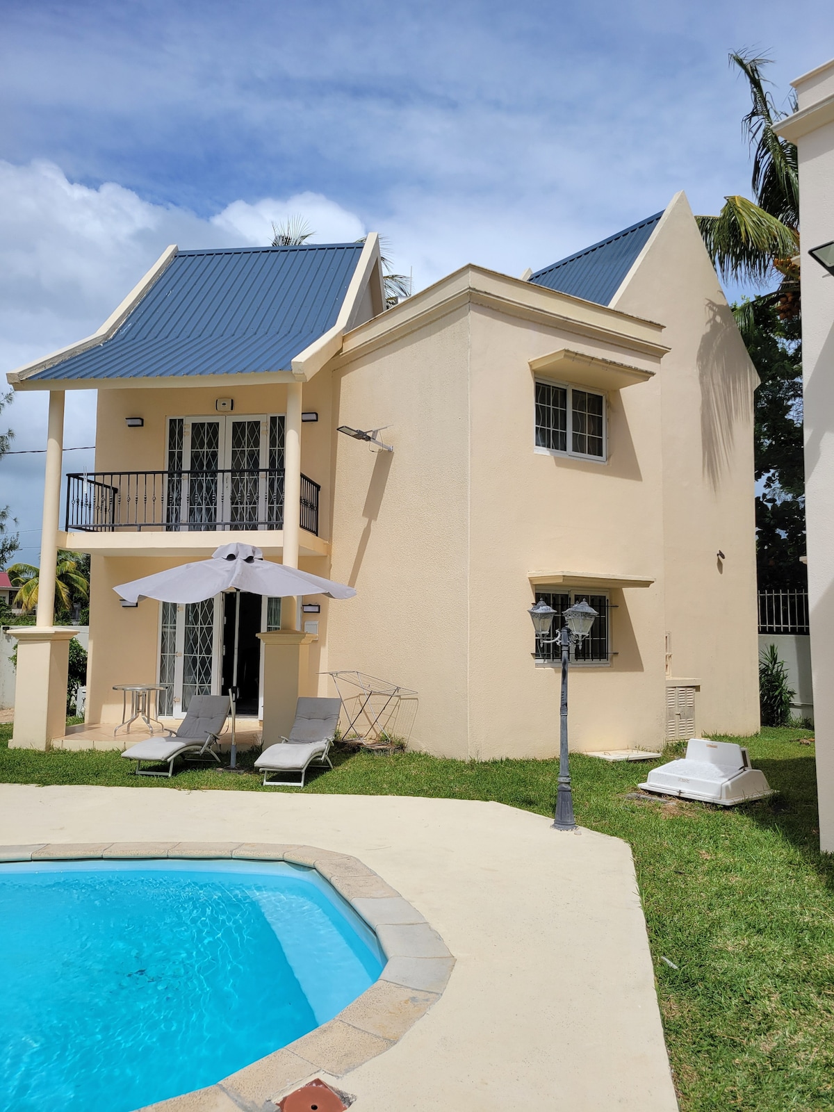 The exterior of the villa is highlighted, featuring a swimming pool in the foreground. Sun loungers are positioned nearby, and the two-story structure presents a combination of architectural elements, including a prominent roof and balcony. Lush greenery surrounds the pool area.