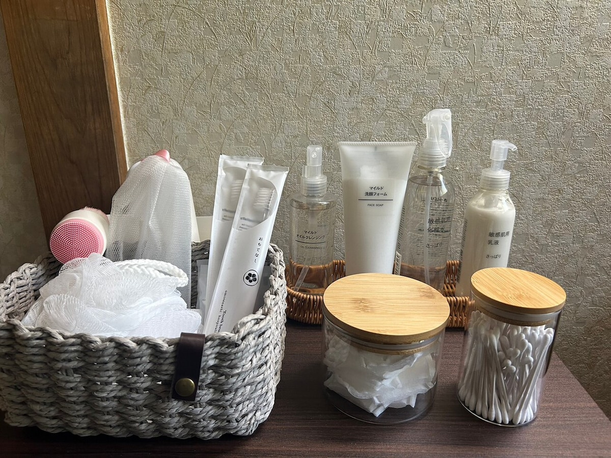 A selection of toiletries is displayed on a wooden surface. A woven basket contains body wash sponges and various skincare products, while two glass jars with bamboo lids hold cotton pads and swabs. Clear bottles contain liquid moisturizers and sprays, organized neatly for convenience.