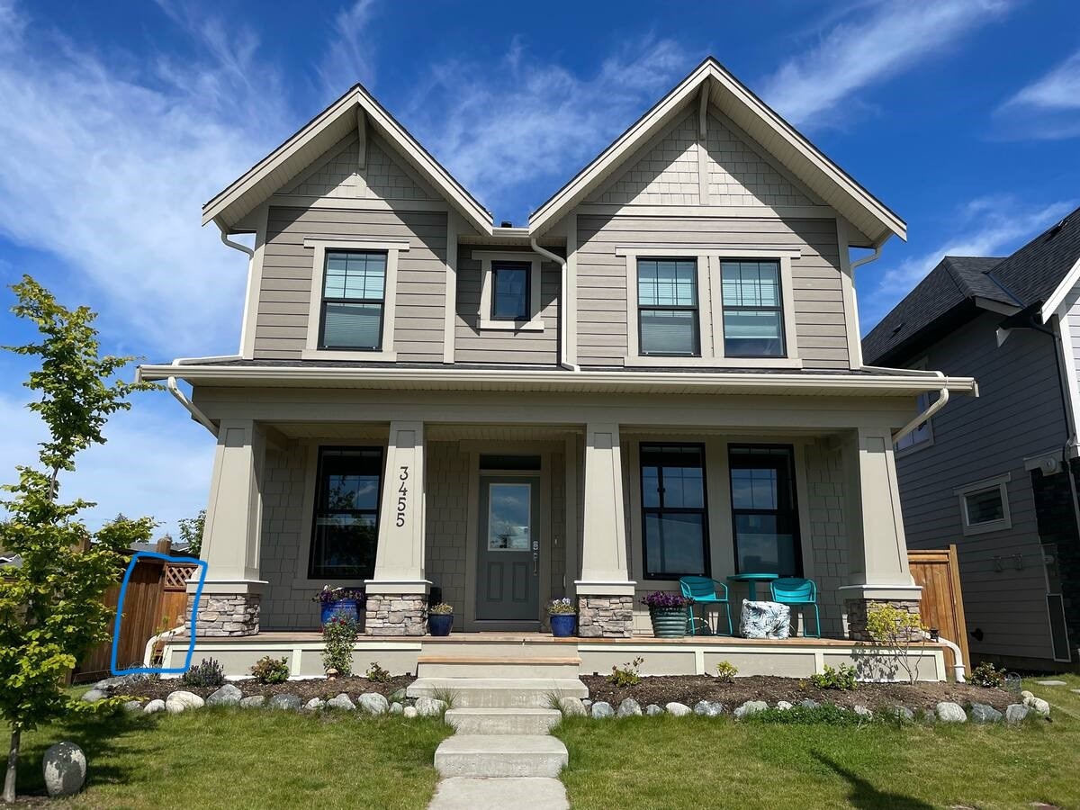 A two-story house with a classic design features a welcoming front porch and a well-maintained garden. Two blue chairs are positioned on the porch, complemented by flower beds and decorative stones lining the pathway to the front door.