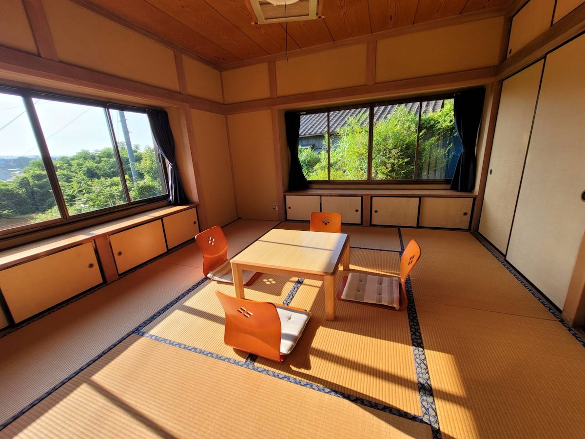 A traditional tatami room features a low table surrounded by four wooden chairs. Large windows provide a view of the greenery outside, filling the space with natural light. Sliding doors offer additional storage and warmth to the inviting design.