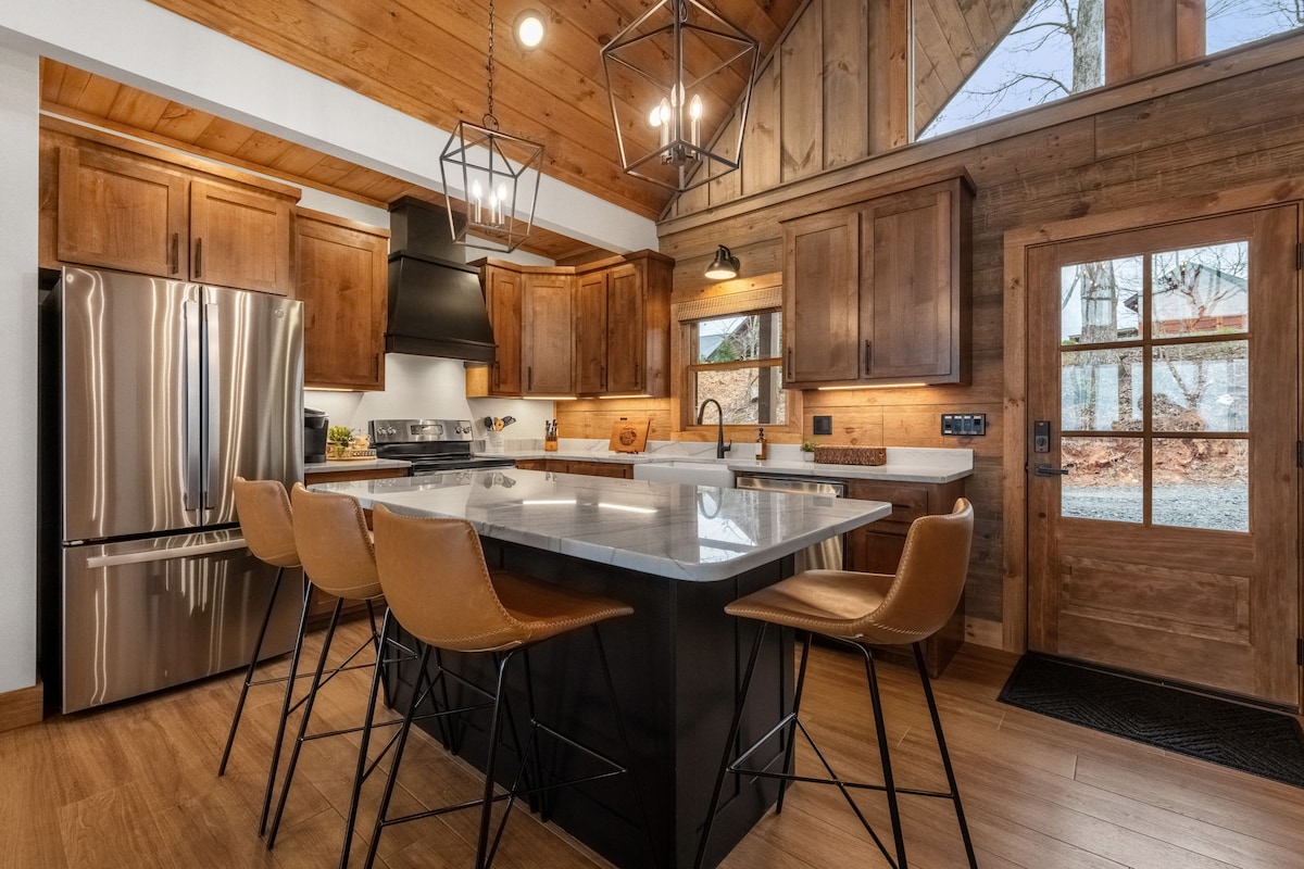 The kitchen features modern wooden cabinetry and stainless steel appliances, including a large refrigerator and a sleek range. A spacious island with a white countertop is complemented by four leather bar stools. Soft lighting enhances the rustic ambiance, while a door leading outside adds natural light.