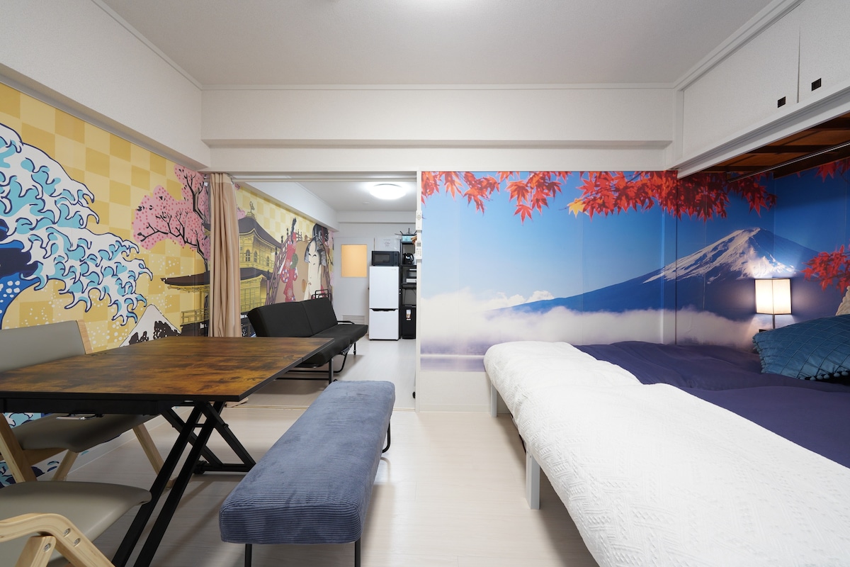 The room features colorful wall murals depicting serene landscapes and cultural motifs. A dining table with chairs is positioned near a bench. Two beds, dressed in white bedding, are separated by a small nightstand, providing a cozy and inviting sleeping area.