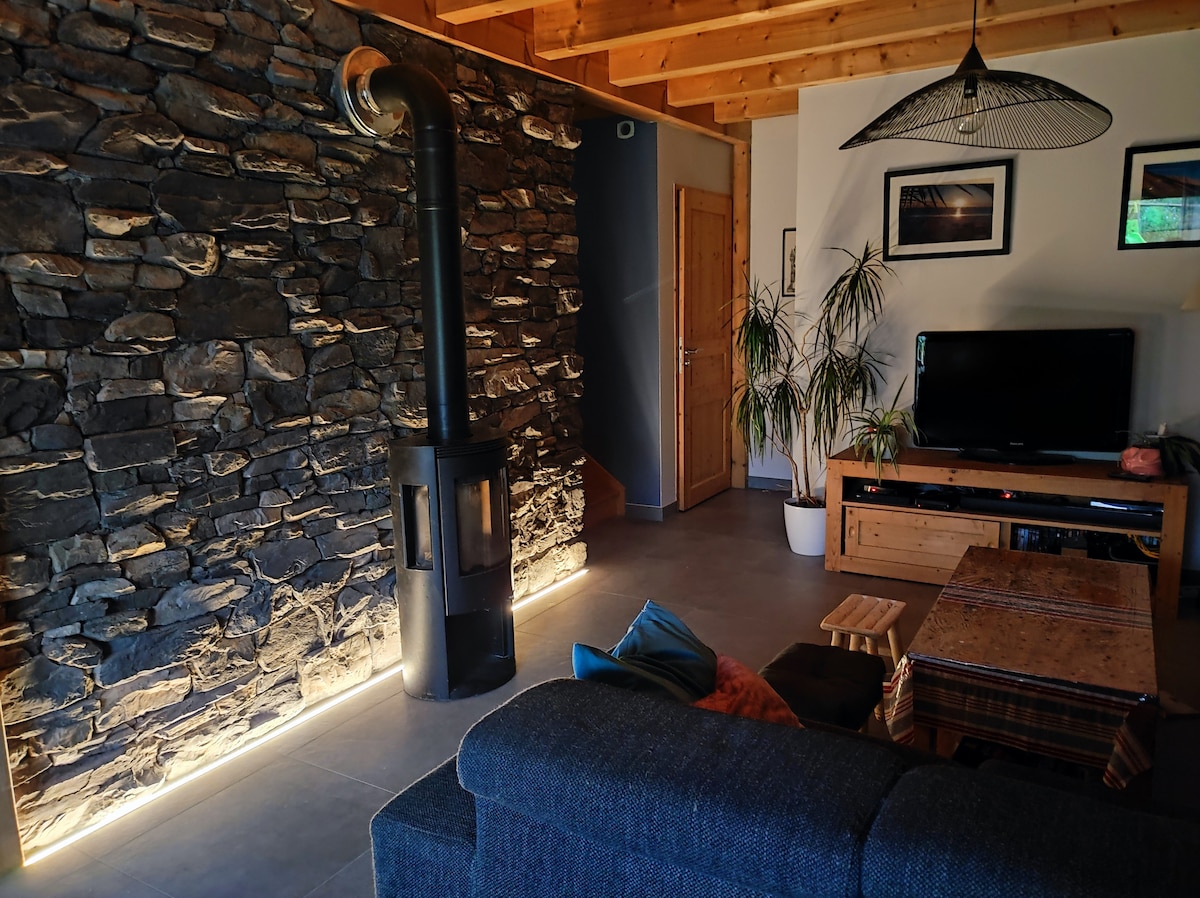 A warm living space is captured, featuring a stone accent wall next to a wood-burning stove. A dark sectional sofa faces a wooden media unit with a television. Soft lighting illuminates the area, creating a cozy ambiance. Potted plants add a touch of greenery.
