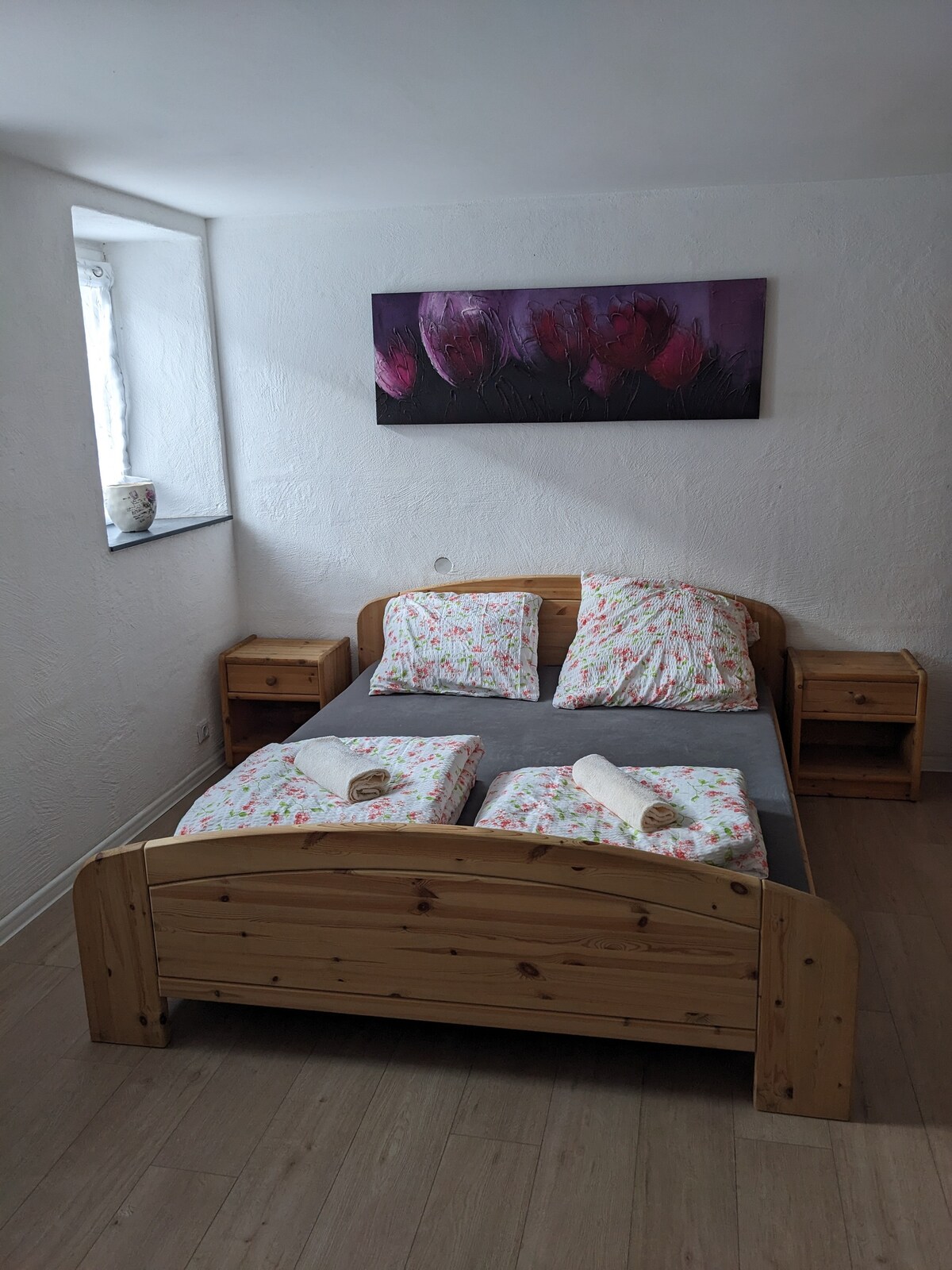 A cozy bedroom is featured with a wooden bed dressed in floral-patterned linens. Two wooden nightstands flank the bed, each topped with rolled towels. A window allows natural light to enter, illuminating the light-colored walls and artwork above the bed.
