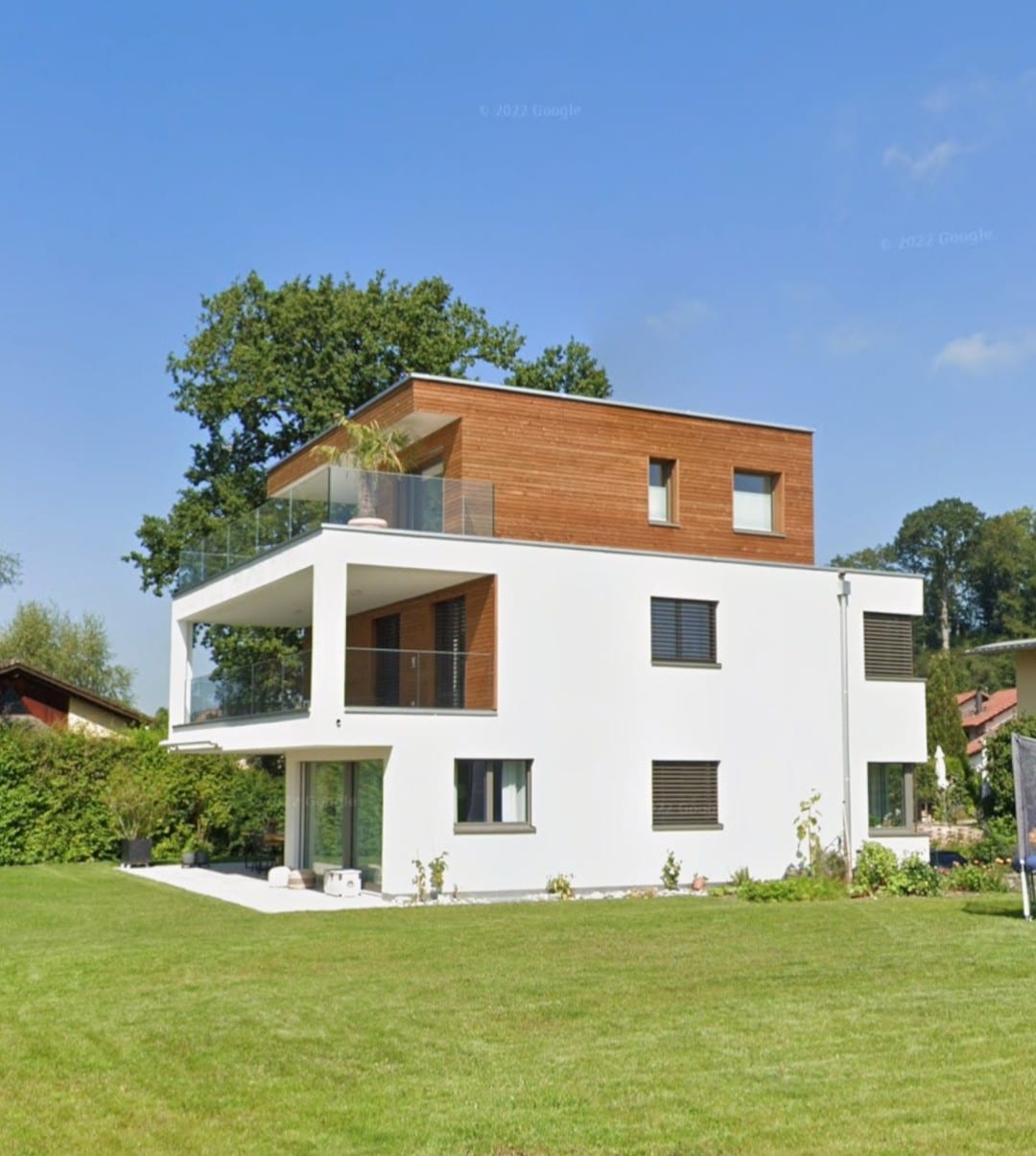 A modern two-story house features a sleek white exterior with wooden accents. Large windows are present, providing natural light to the interiors. A spacious lawn surrounds the property, contributing to a peaceful outdoor environment. A terrace is visible on the upper level, enhancing the design.