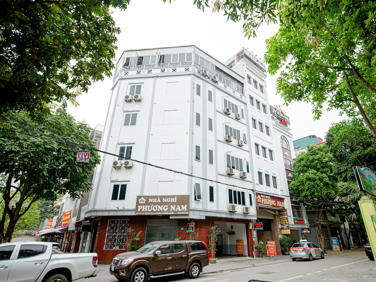 The exterior of Phuong Nam Hotel is captured, showcasing a multi-story building with a white facade and large windows. Air conditioning units are visible on the walls. Lush greenery from surrounding trees adds a natural touch to the setting, with nearby shops and vehicles also in view.