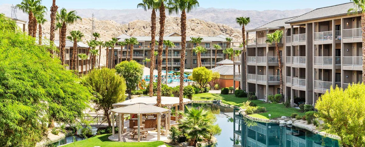 The view captures a well-kept resort surrounded by palm trees and vibrant greenery. Gazebos are set by a serene water feature that reflects the surrounding landscape. Multiple floors of the buildings are visible, offering a glimpse of outdoor swimming areas and relaxation spaces.