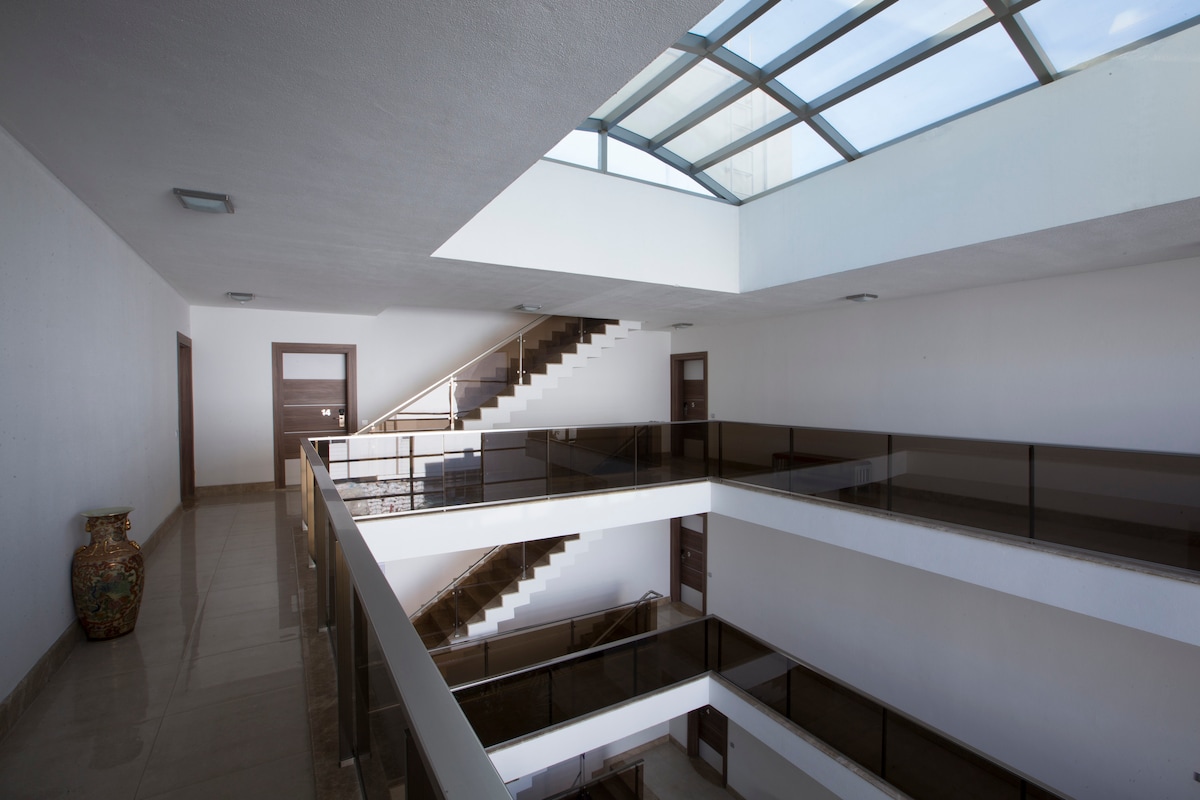 A spacious atrium showcases a modern design with multiple levels connected by elegant staircases. Natural light filters through a large skylight, illuminating the white walls and polished tile flooring. Glass railings enhance visibility while decorative elements are present in the corners.