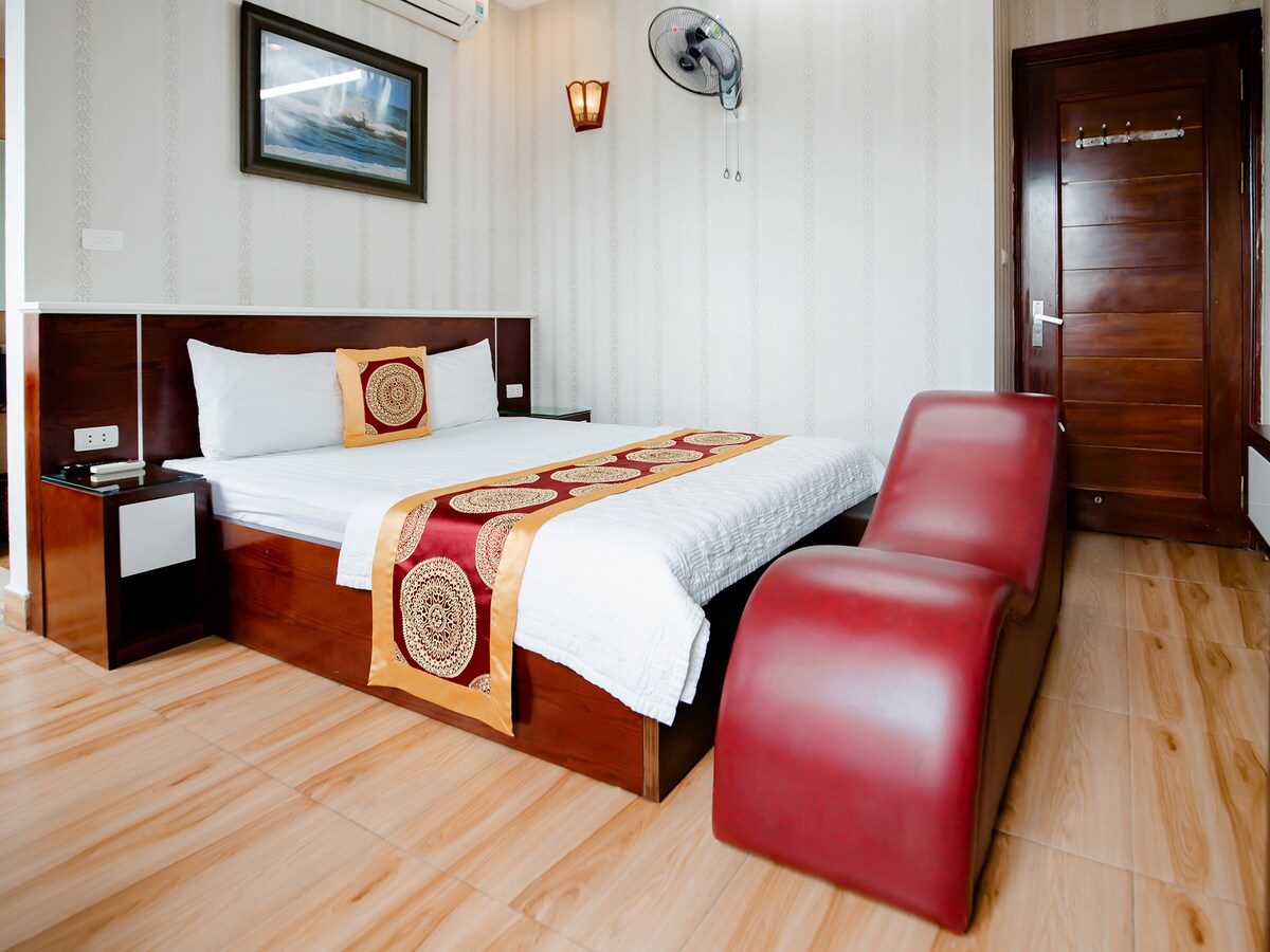 A private room features a spacious bed dressed in white linens, complemented by a decorative throw. A red lounge chair is positioned nearby, while natural light filters through the window. The wooden door and wall art accentuate the room's design.