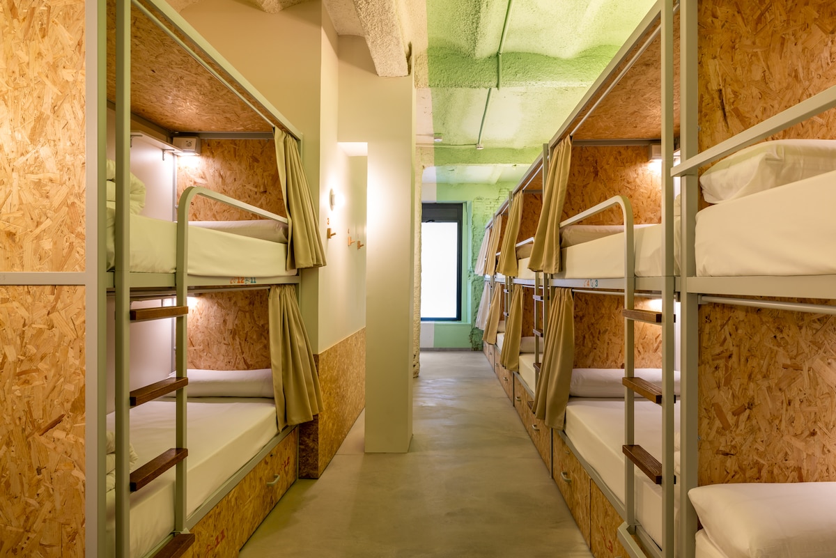 A well-lit dormitory features multiple bunk beds, each equipped with individual lights and curtains for privacy. Natural light is visible entering through a large window at the end of the hallway, illuminating the minimalist design and the warm tones of the wood paneling.