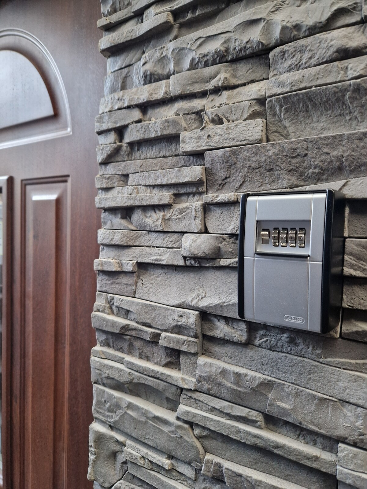 A close-up view of a stone wall adjacent to a wooden door, featuring a sleek electronic lockbox. The textured stone has different shades of grey, providing a modern contrast to the rich brown of the door.