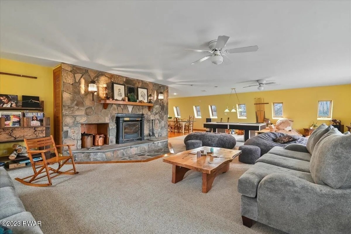 A spacious living area features a stone fireplace with wood storage and a large, comfortable sectional sofa. Natural light floods in through multiple windows, illuminating the warm yellow walls. A wooden table and chairs are visible in the background, alongside a game table.