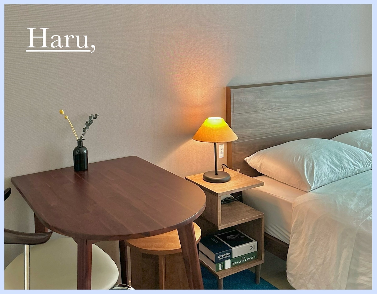 A small wooden table is positioned adjacent to a softly lit lamp on a nightstand. A spacious bed is covered in white linens, complemented by light-colored walls. A decorative vase with minimal greenery adds a touch of elegance to the space.