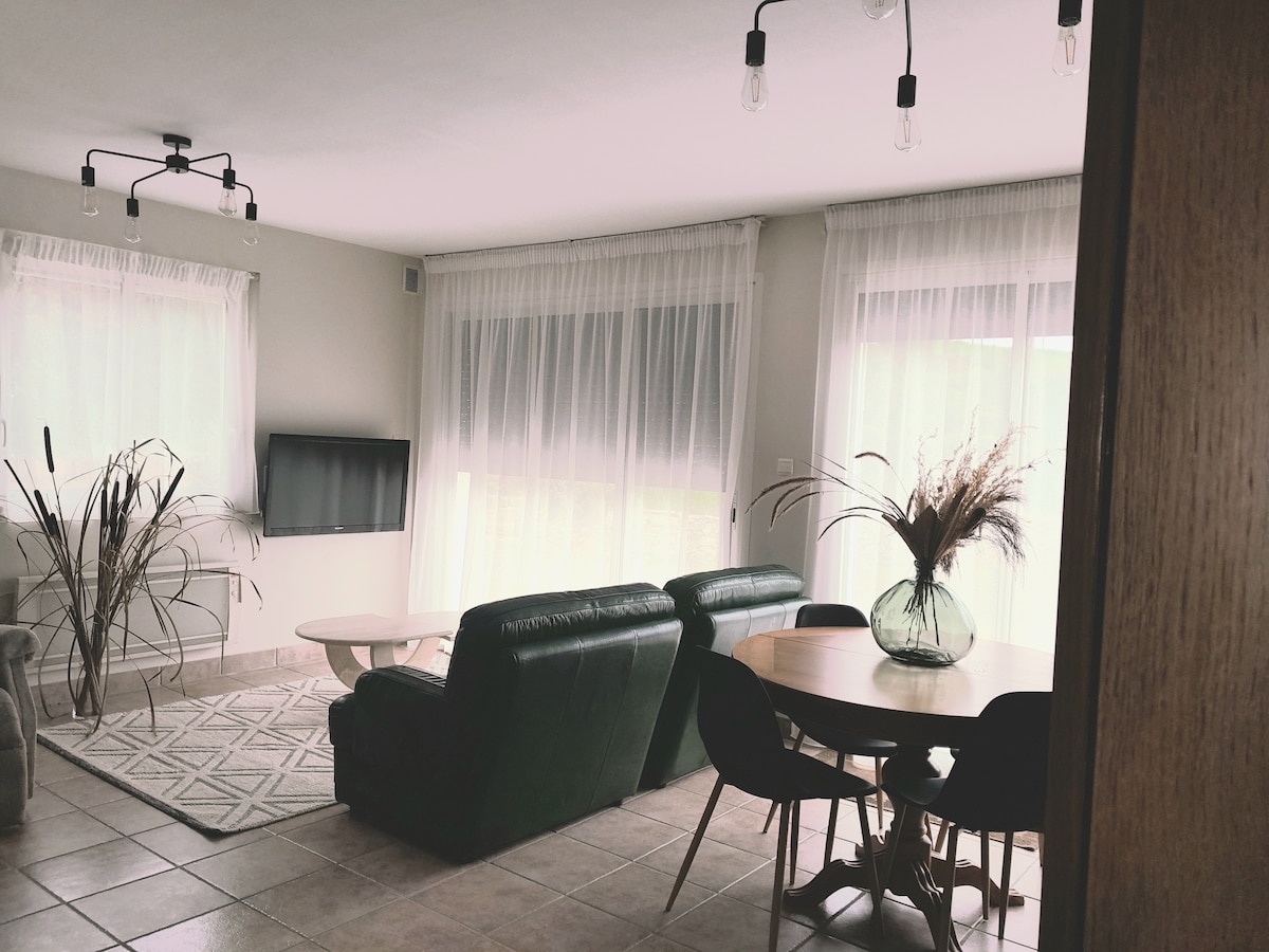 A modest living area is presented with large windows dressed in sheer curtains, allowing natural light to fill the space. A sleek television is mounted on the wall, while a round dining table surrounded by chairs complements two comfortable sofas. A minimalist vase with dried plants decorates the table.