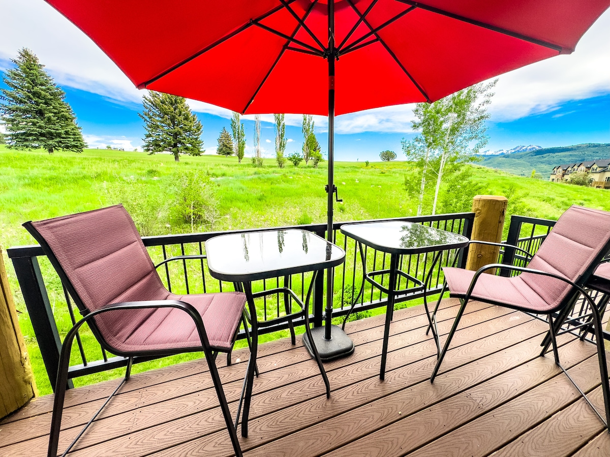 A balcony is presented with a red umbrella providing shade over a black table and two pink chairs. Lush green fields and trees are visible in the background, framed by a clear blue sky and distant mountains.