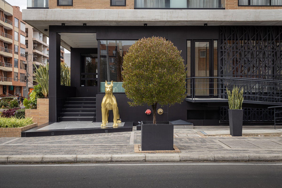 The exterior of the building is highlighted by a golden dog statue positioned at the entrance, surrounded by neatly trimmed greenery and modern planters. A ramp provides accessible entry, while the contemporary facade features a mix of dark tones and natural elements.