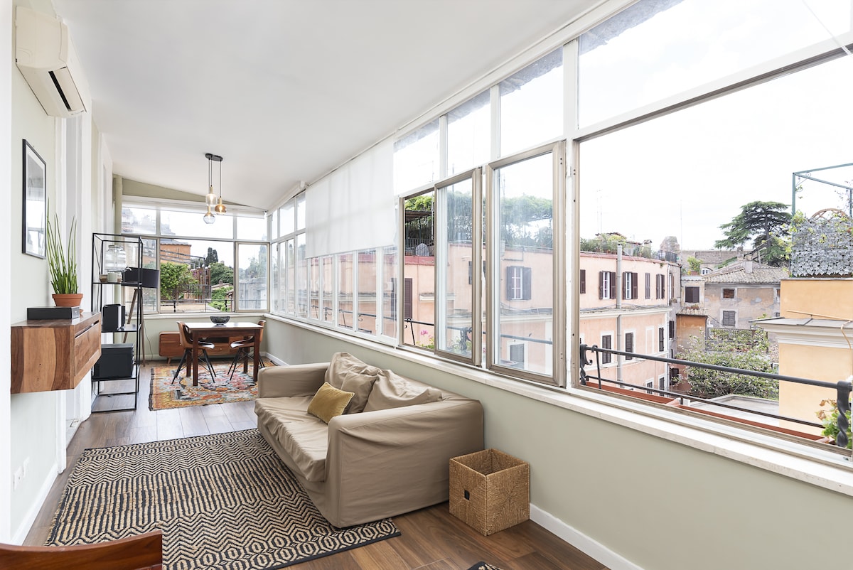 A sunroom features large windows that provide ample natural light and offer views of surrounding buildings and greenery. A comfortable sofa is positioned near the windows, accompanied by a dining table for four. Decorative elements, like a patterned rug, enhance the interior space.