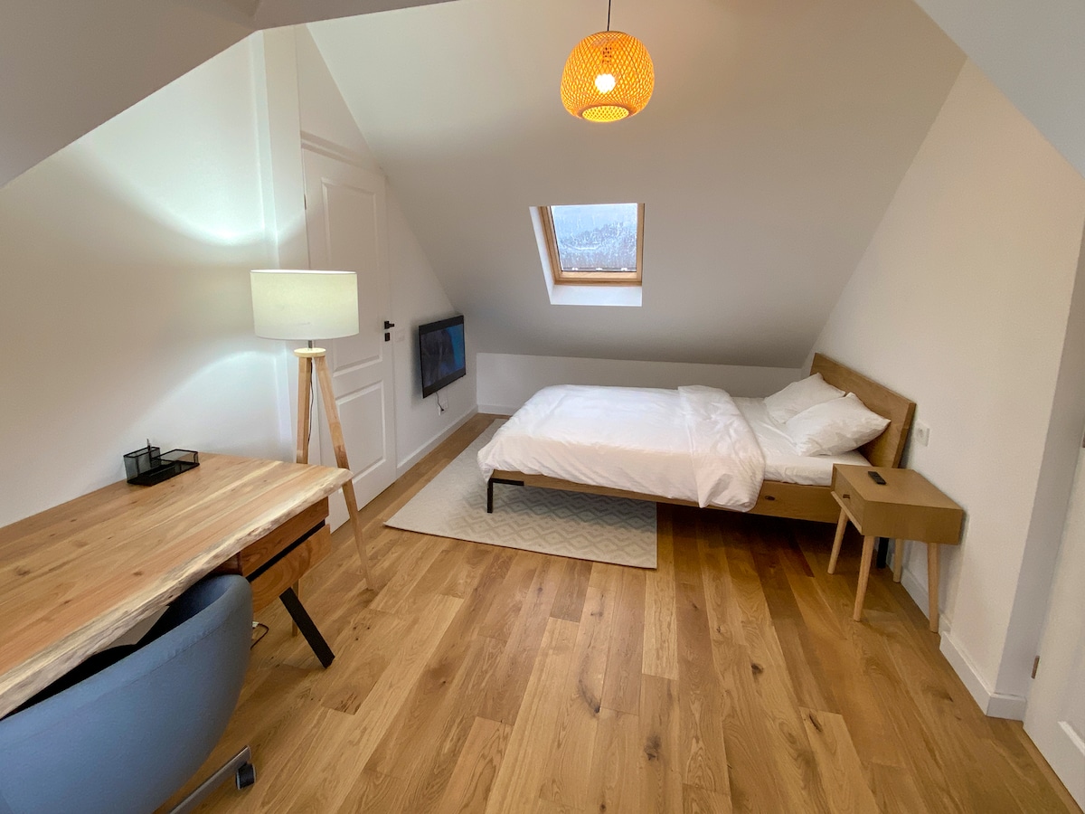 A light-filled attic room features a spacious bed dressed in white linens, complemented by wooden furniture. A desk with a blue chair is positioned near a window, while a modern lamp and a wall-mounted television enhance the functionality of the space.