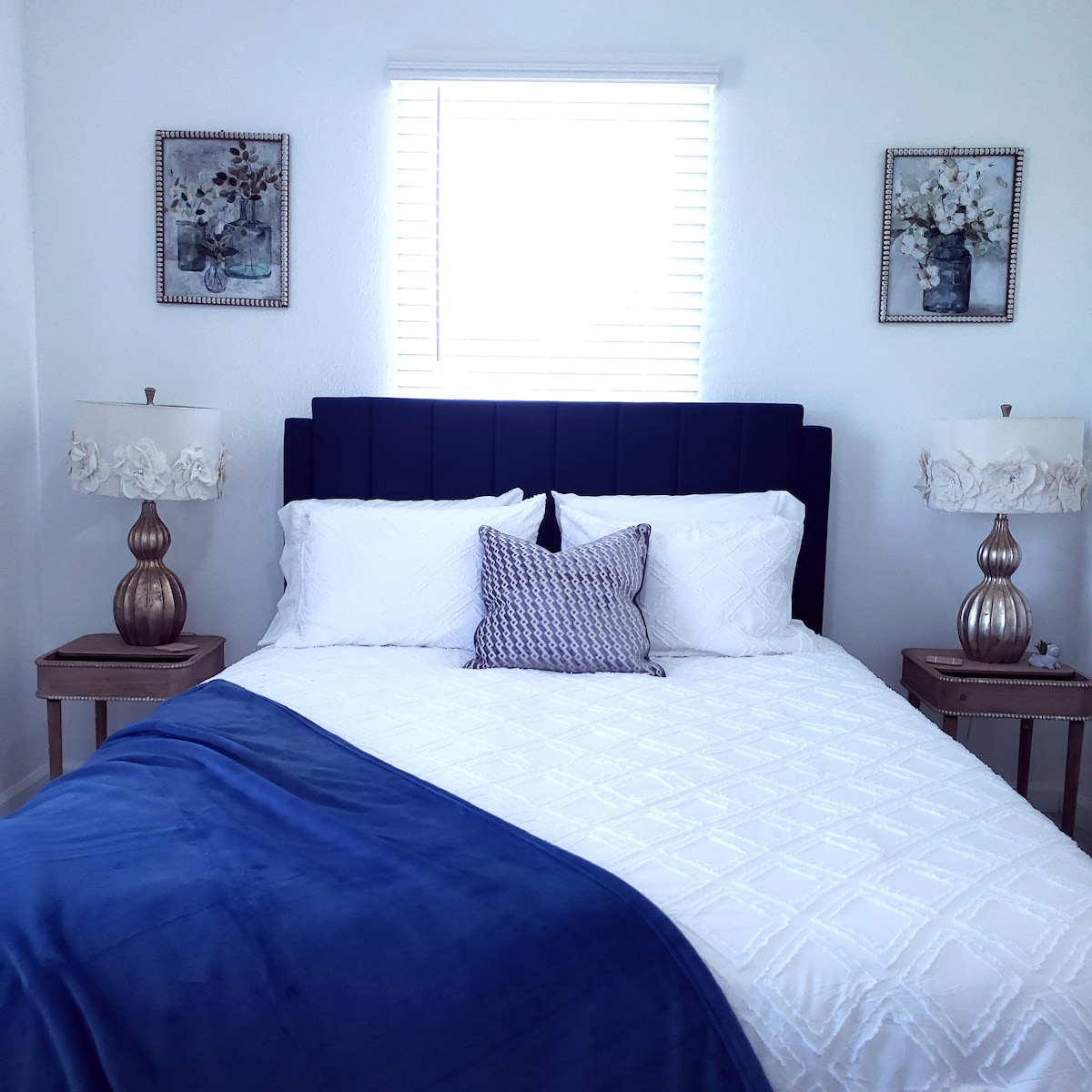 A spacious bed is adorned with white linens and a textured navy throw blanket. Coordinating pillows add a subtle touch of color. Two bedside lamps with floral designs are placed on stylish nightstands beside the bed. Natural light enters through a window covered by horizontal blinds.