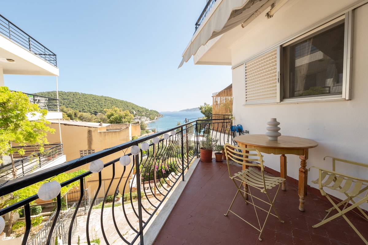 A balcony is equipped with a round wooden table and two iron chairs, offering a space for relaxation. Decorative lanterns add a charming touch. The view includes lush greenery and a clear sea, with the coastline visible in the distance.
